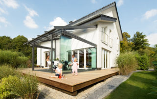 Weißes Einfamilienhaus mit hölzerner Terrasse und Glashaus, eltern und zwei spielende Kleinkinder