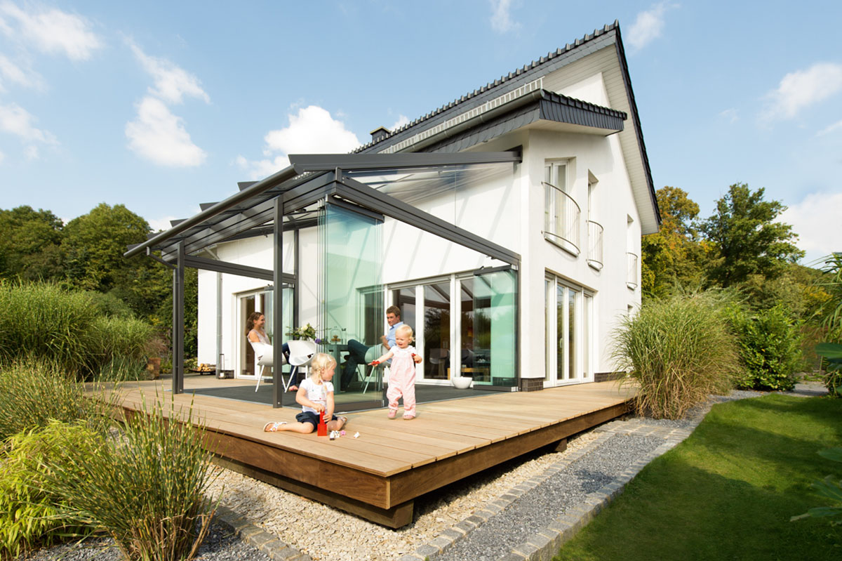 Weißes Einfamilienhaus mit hölzerner Terrasse und Glashaus, eltern und zwei spielende Kleinkinder